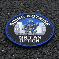 A circular embroidered patch with a blue border and the text 'DOING NOTHING ISN'T AN OPTION' around a graphic of a tactical officer in front of city buildings. The patch is placed on tarmac